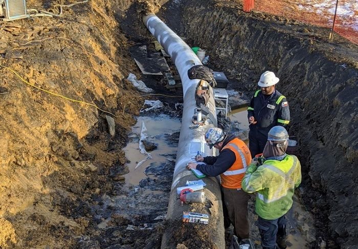 Field engineers and technicians in high-visibility gear and hardhats conducting non-destructive testing (NDT) inspection on an exposed pipeline section inside an excavation trench.